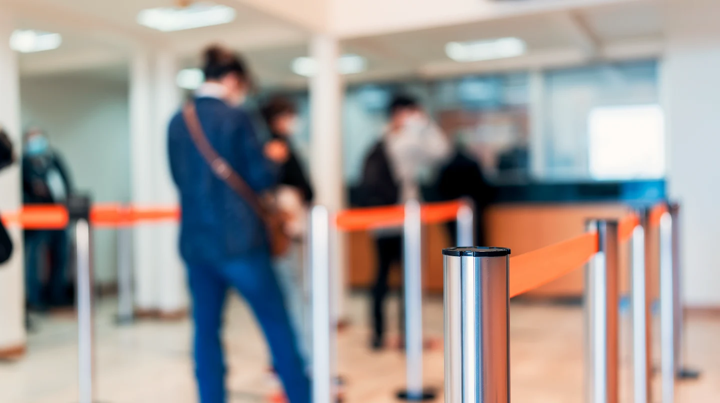 Customers waiting in line at a bank branch behind orange retractable rope stanchions leading to the teller counters.